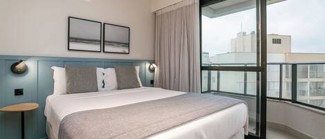 A modern hotel room with a large bed, a small side table, a large window with curtains, and a city view.