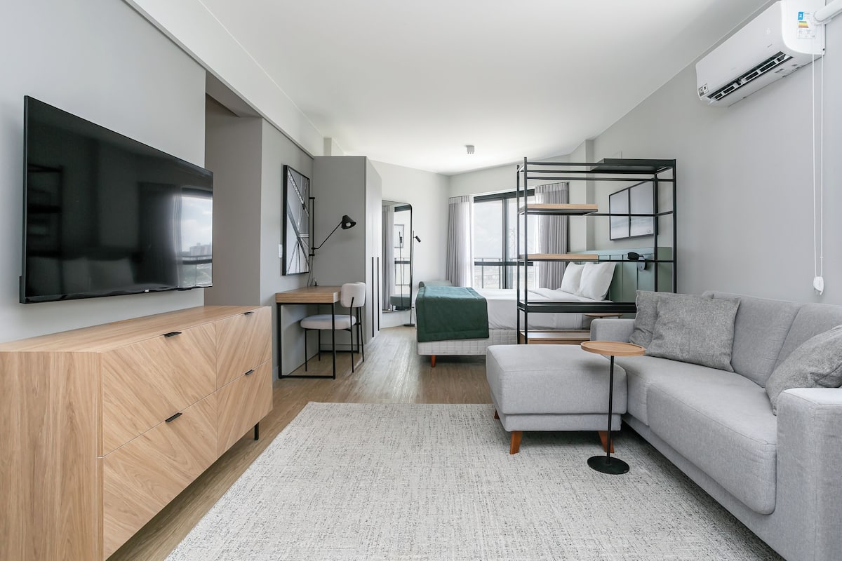 A modern living room with a flat-screen TV, a wooden cabinet, a grey sofa, a small round table, and a bunk bed with a green blanket.