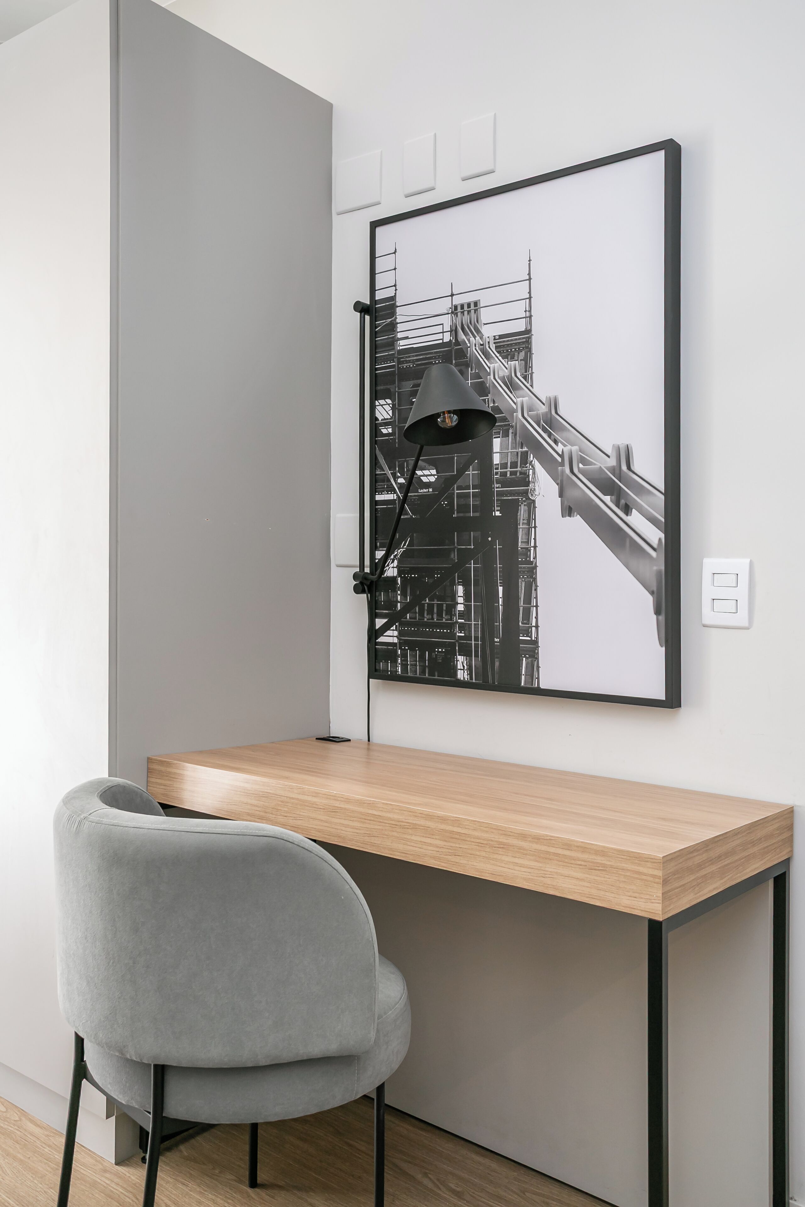 A modern desk with a grey chair, a framed black and white photograph on the wall, and a black desk lamp.