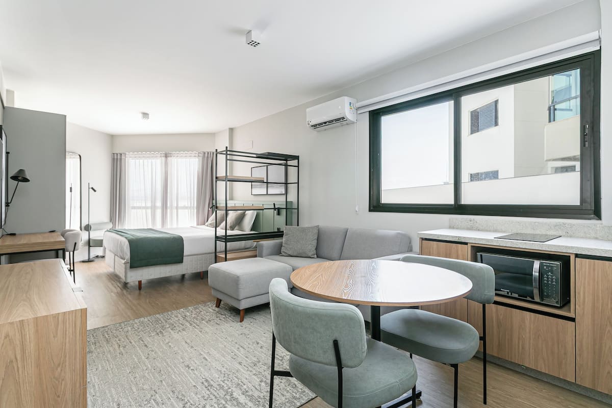 A modern, well-lit room with a bed, sofa, dining table, and kitchenette.