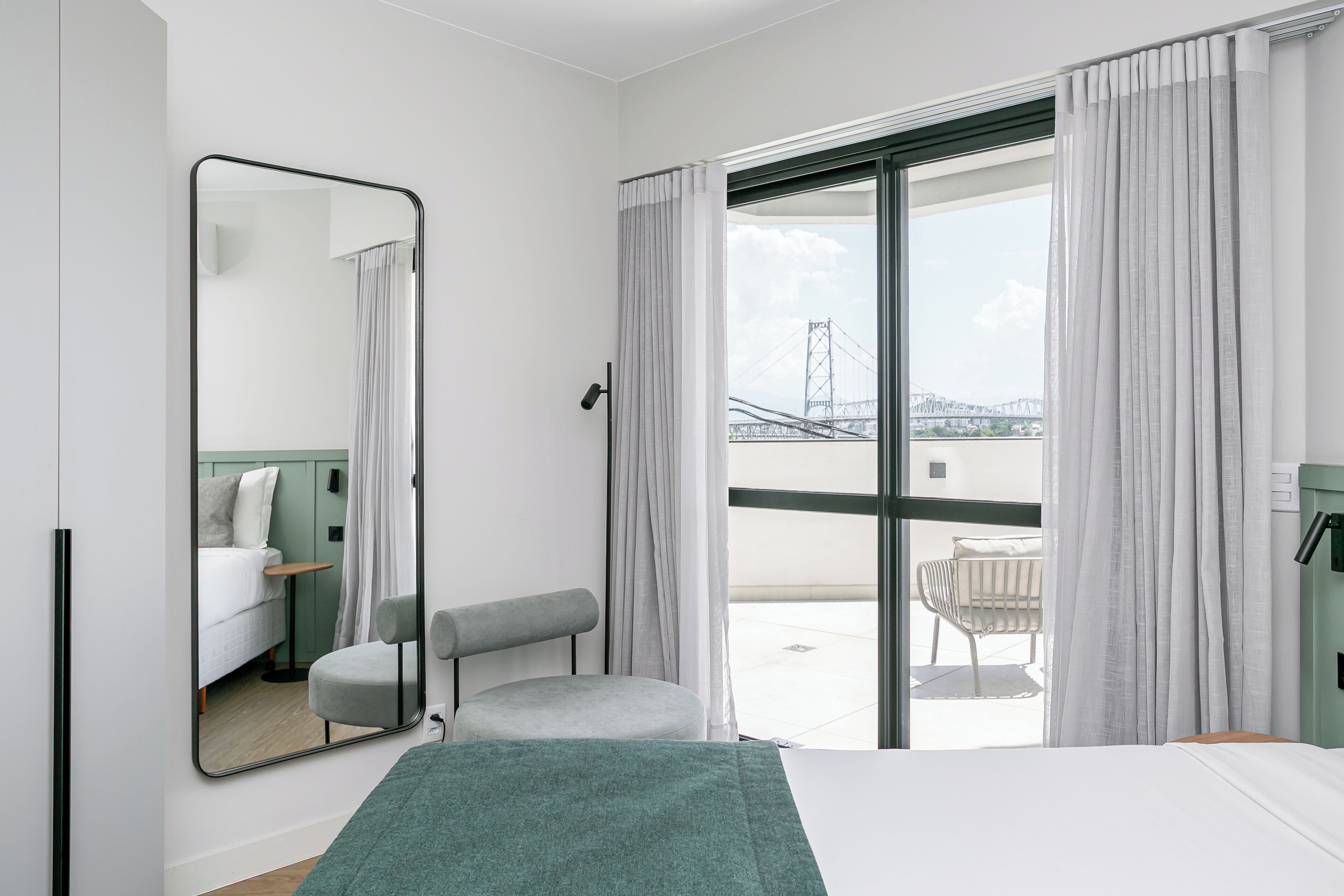 A modern hotel room with a large bed, a seating area with a chair and a small table, a large mirror, and a view of a bridge through the window.