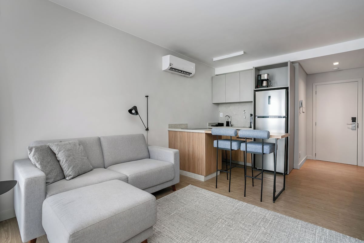 A modern apartment with a grey sofa, a kitchen area with a fridge and a coffee machine, and a small bar area.