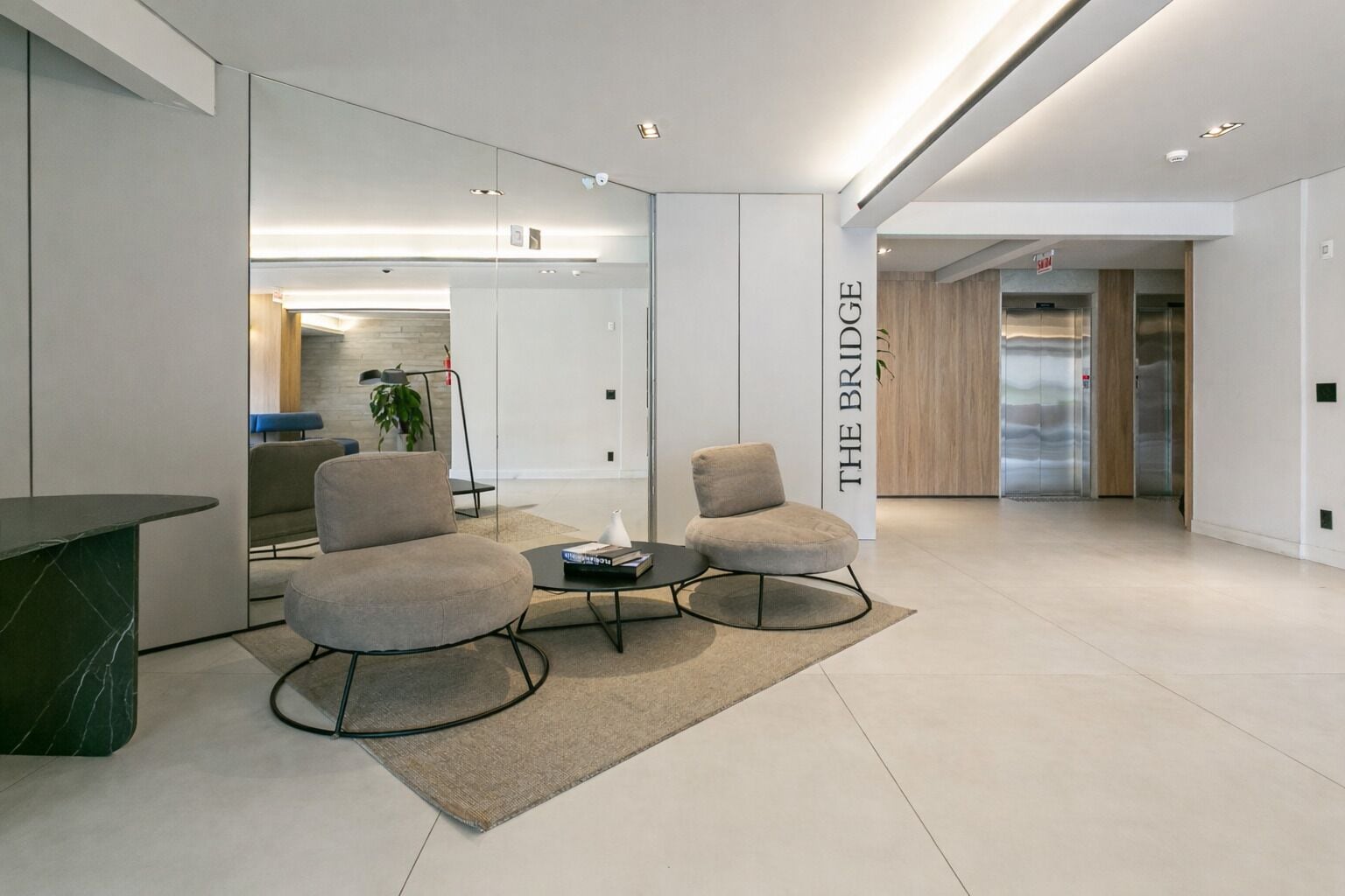 A modern, well-lit lobby with a marble reception desk, two grey armchairs, a small coffee table, and a wall-mounted sign reading "THE BRIDGE".