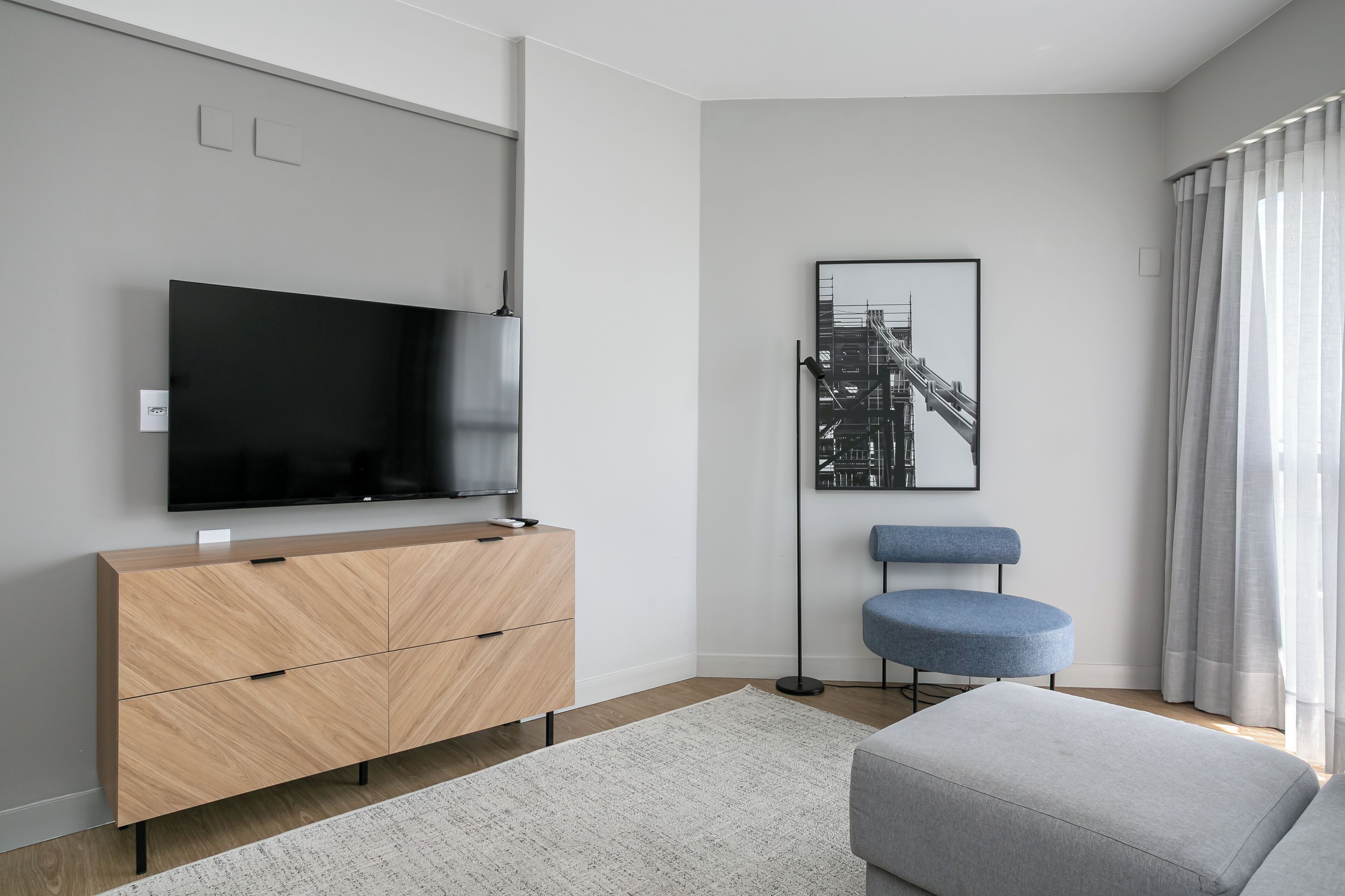 Ein modernes Wohnzimmer mit einem an der Wand montierten Flachbildfernseher, einem hellen Holzschrank, einem blauen Sessel und einem grauen Hocker.