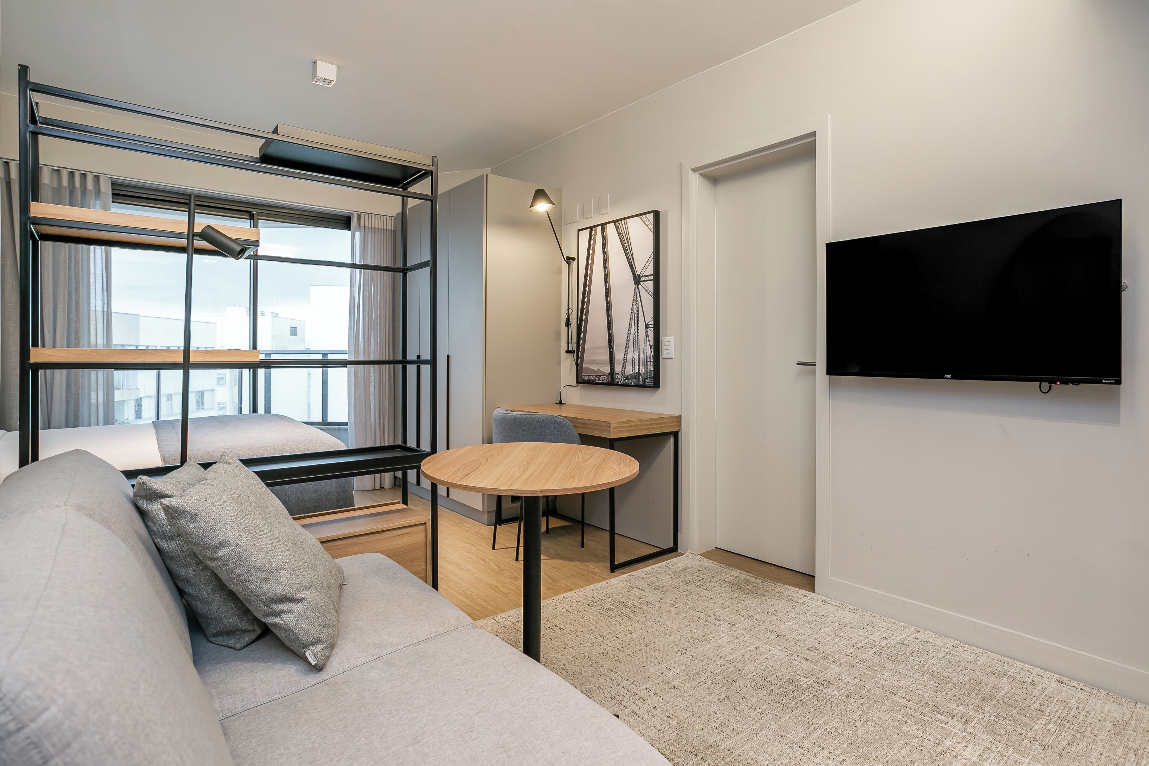 Un espace de vie moderne et compact, comprenant un lit superposé, un canapé, une petite table ronde et un téléviseur à écran plat fixé au mur.