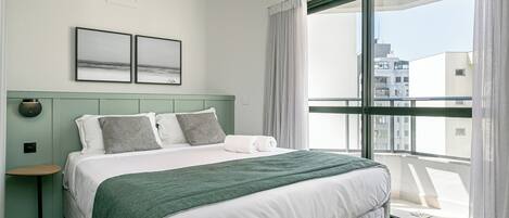 A modern hotel room with a large bed, a view of the city through a large window, and a small side table.