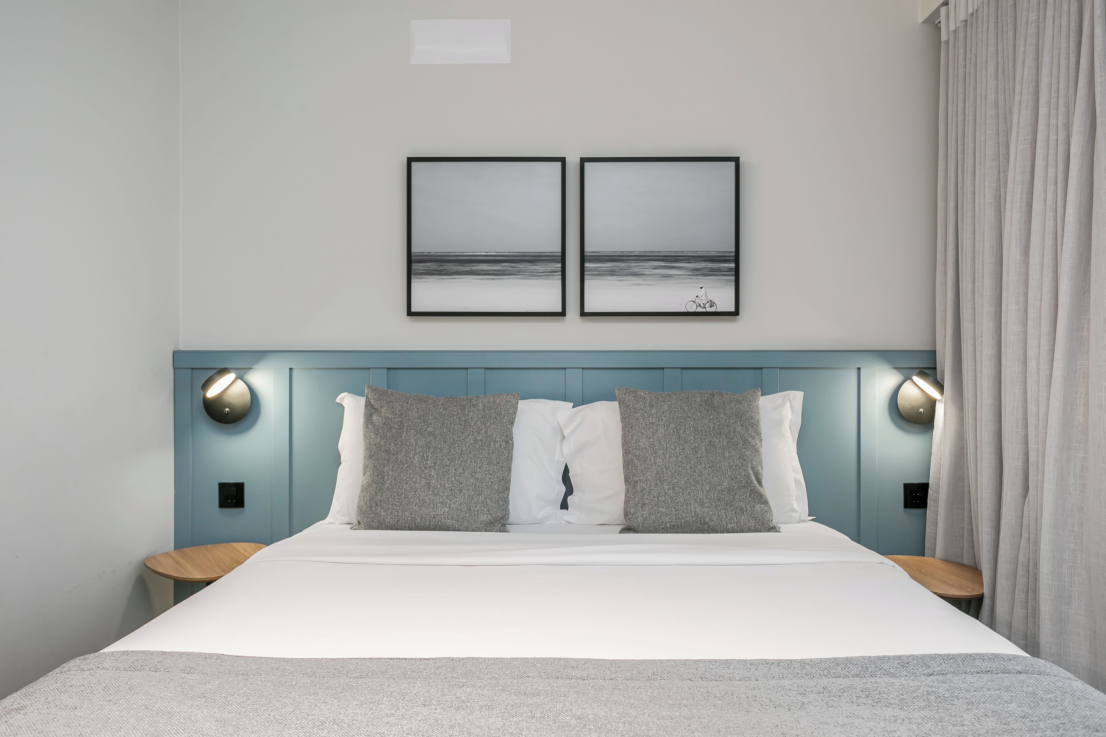 A neatly made bed with a white and gray color scheme, two framed pictures on the wall, and a blue headboard.