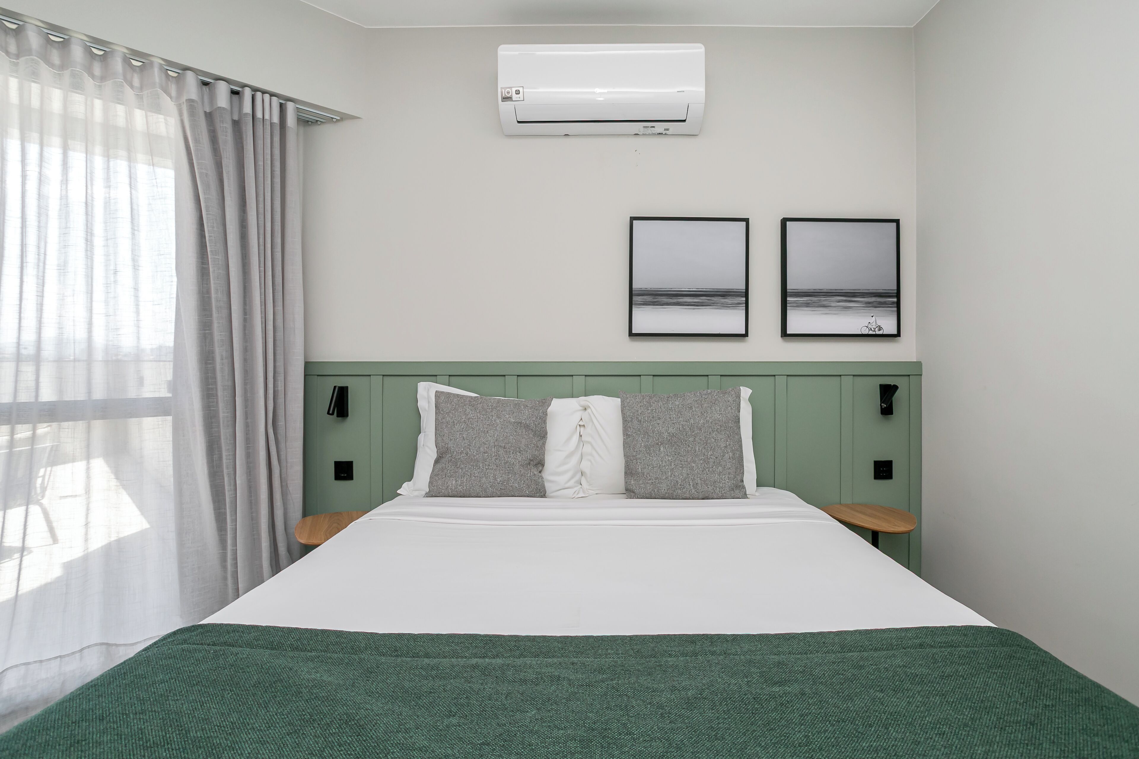 Une chambre d’hôtel bien rangée, avec un lit, une fenêtre avec des rideaux, un appareil de climatisation, deux tableaux encadrés et une tête de lit verte.