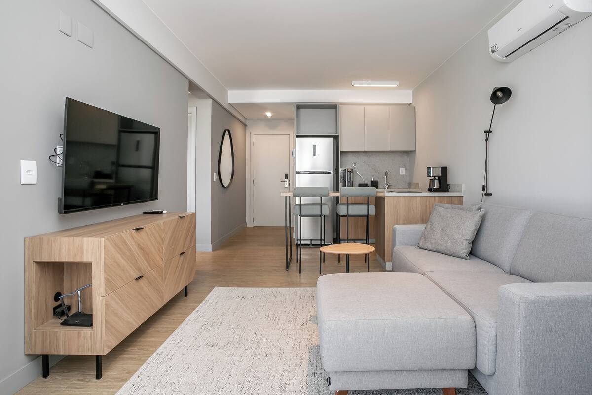 A modern living room with a grey sofa, a TV mounted on the wall, a wooden cabinet, and a kitchen area in the background.