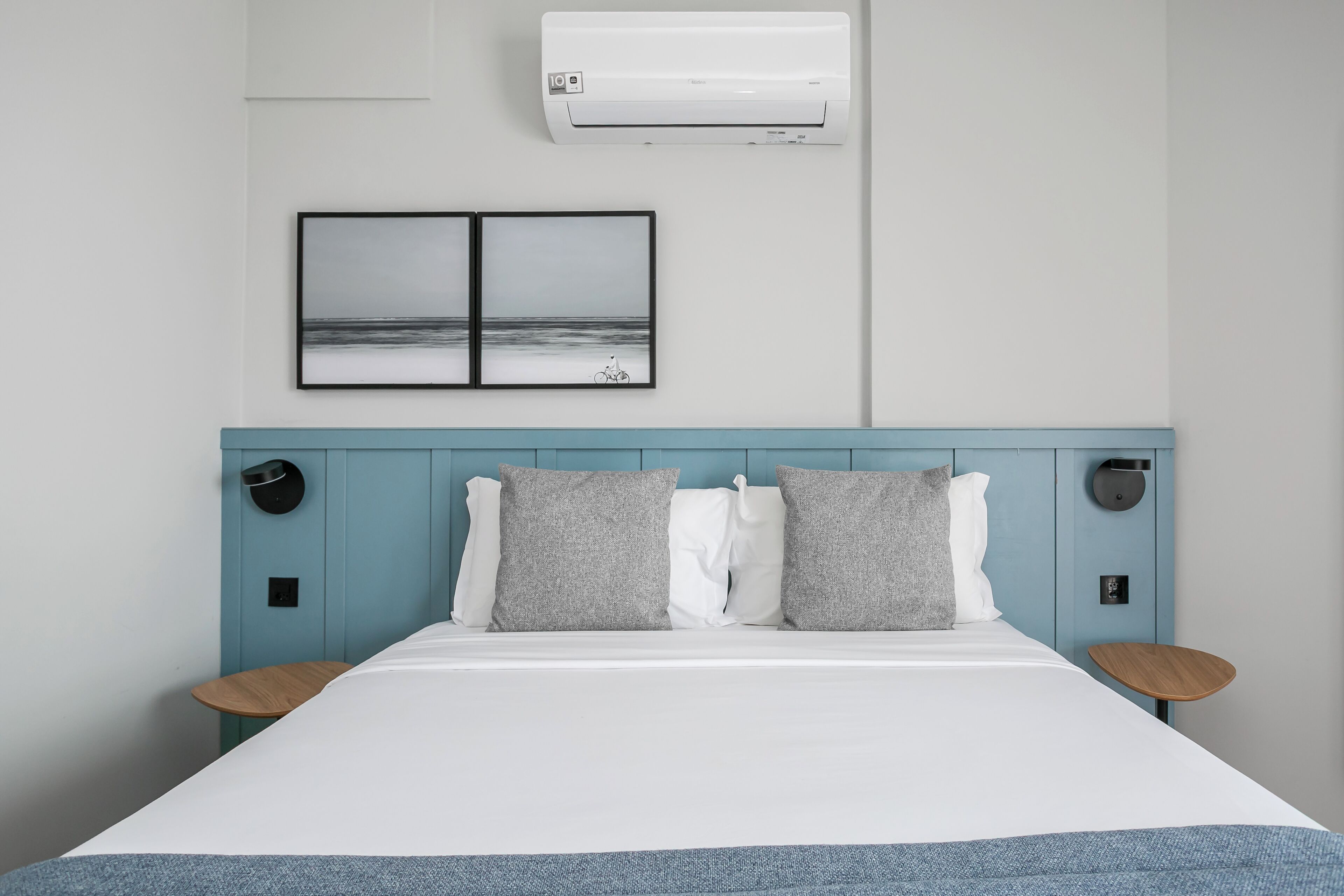 A neatly made bed with a white duvet, two grey pillows, and a blue headboard. There is a wall-mounted air conditioner above the bed and a framed picture of a beach scene below it.