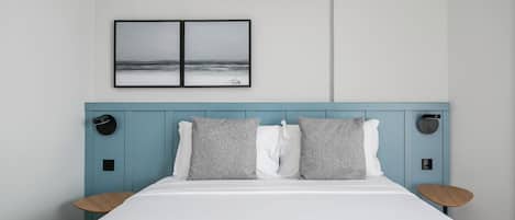 A neatly made bed with a white duvet, two grey pillows, and a blue headboard. There is a wall-mounted air conditioner above the bed and a framed picture of a beach scene below it.