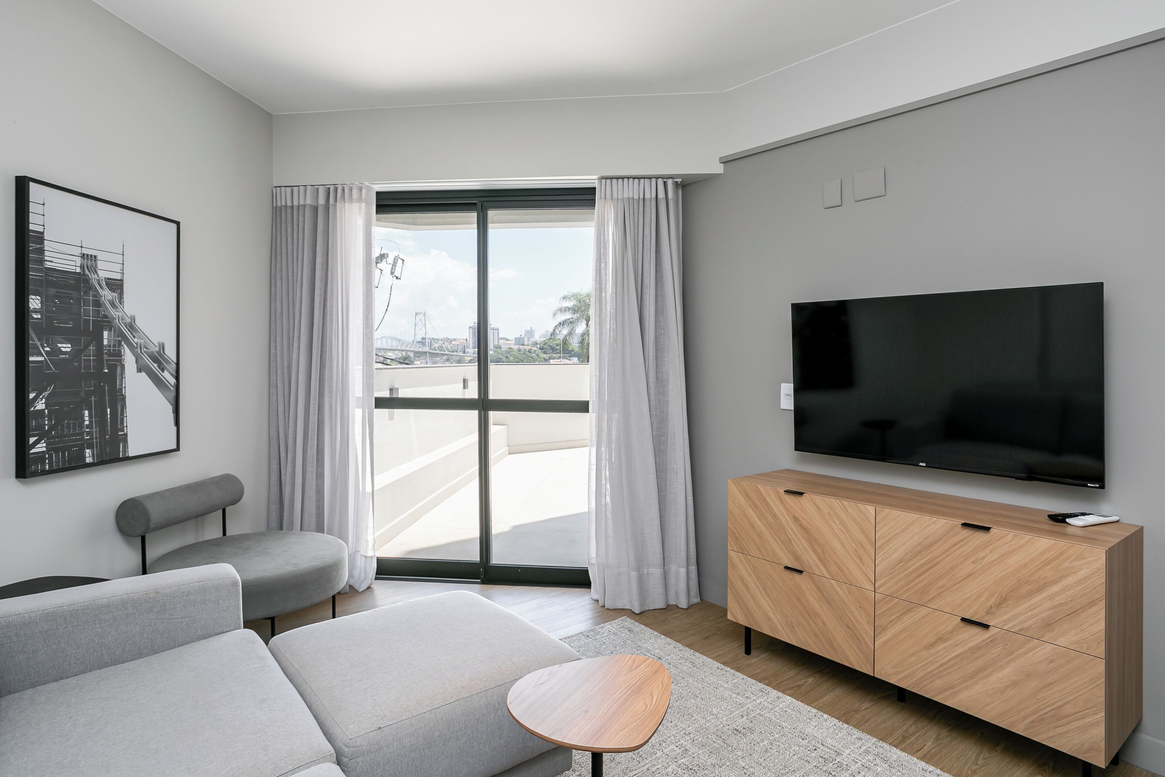 A modern living room with a large flat-screen TV, grey sofas, a wooden cabinet, and a large window with curtains.