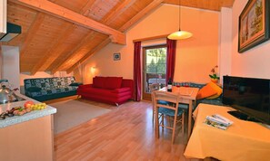 A cozy loft-style room with a wooden ceiling, a red sofa, a dining table with chairs, a TV, and a kitchen area with a kettle and fruit. - Apartment 'Bergblick' with Mountain View, Shared Terrace and Wi-Fi (St. Maddalena)