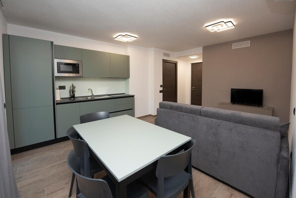 A modern kitchen with a microwave, a dining table with chairs, a gray sofa, and a TV on a stand.