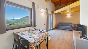 A dining area with a table set for two, a sofa, and a television in a room with wooden beams and large windows. - Suly Apartments 3 – Modern Accessible Stay with Mountain Views in Saint-Vincent (Cillian)