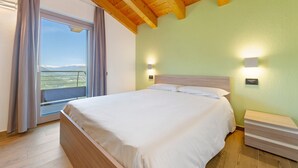 A bedroom with a bed, a nightstand, a window with curtains, and a view of the outdoors. - Suly Apartments 3 – Modern Accessible Stay with Mountain Views in Saint-Vincent (Cillian)