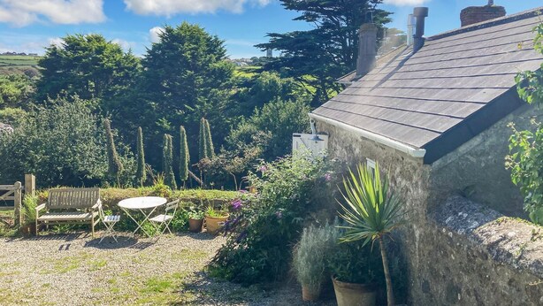 A rustic stone house with a sloped roof, surrounded by a garden with various plants and a wooden bench. - The Old Studio (Penzance)