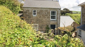A stone house with two windows and a door, surrounded by lush greenery. - The Old Studio (Penzance)