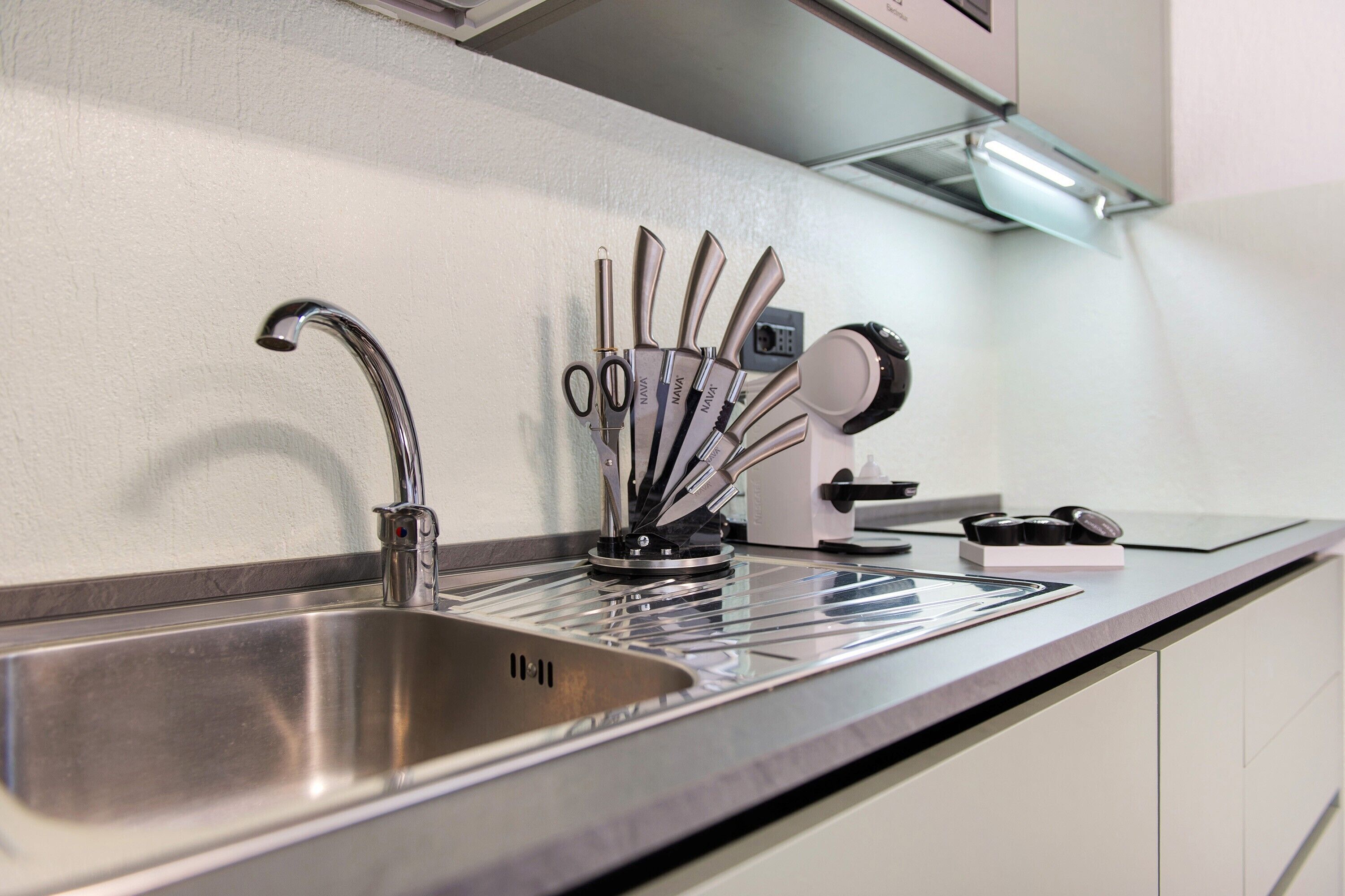 Un évier de cuisine en acier inoxydable, doté d’un robinet moderne, un bloc à couteaux contenant différents types de couteaux et une machine à café posée sur le plan de travail.