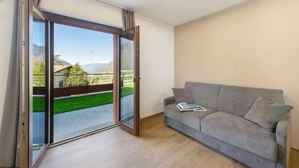 A room with a sliding glass door leading to a balcony, a grey sofa with a book, and a modern light fixture. - Suly Apartments 1 - cir0082 (Cillian)
