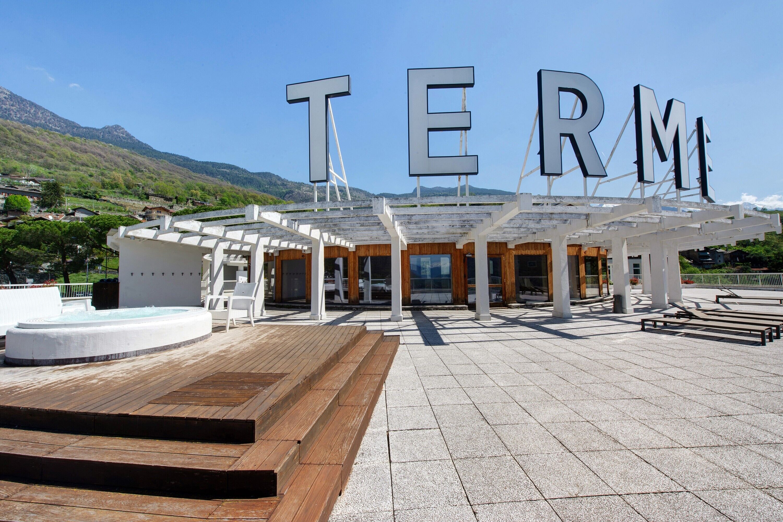 Un bâtiment moderne dont la façade est ornée de grandes lettres formant le mot « TERM », avec une terrasse en bois et un espace piscine.
