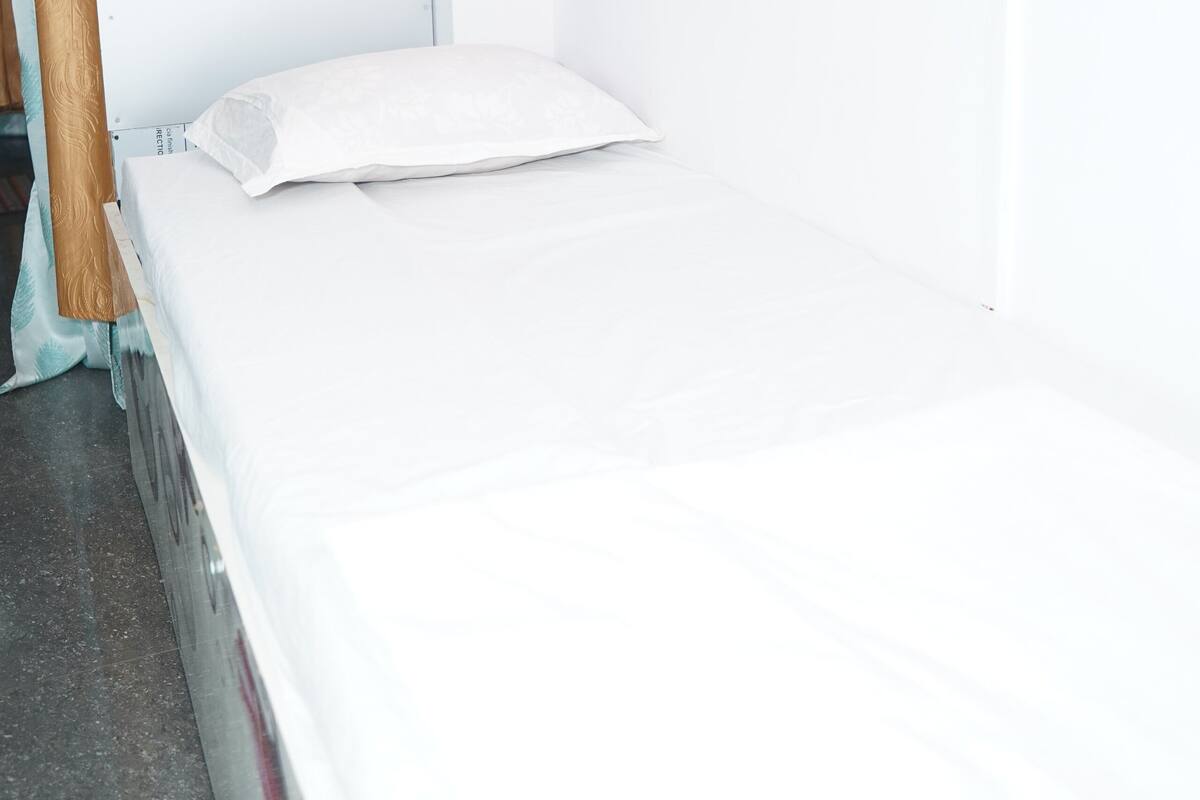 Una habitación pequeña con cama individual, cabecera de madera, ropa de cama blanca y pared lisa de color blanco.
