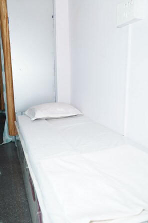 A small, single-bed room with a white mattress and pillow, a wooden headboard, and a white wall with a light switch. - Meera Guest House (Bhagalpur)