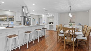 Una cocina moderna con armarios blancos, una isla central con taburetes y un comedor con sillas de madera y una mesa.
