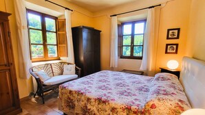 Ein Schlafzimmer mit Bett, Stuhl, Kleiderschrank und zwei verhangenen Fenstern.