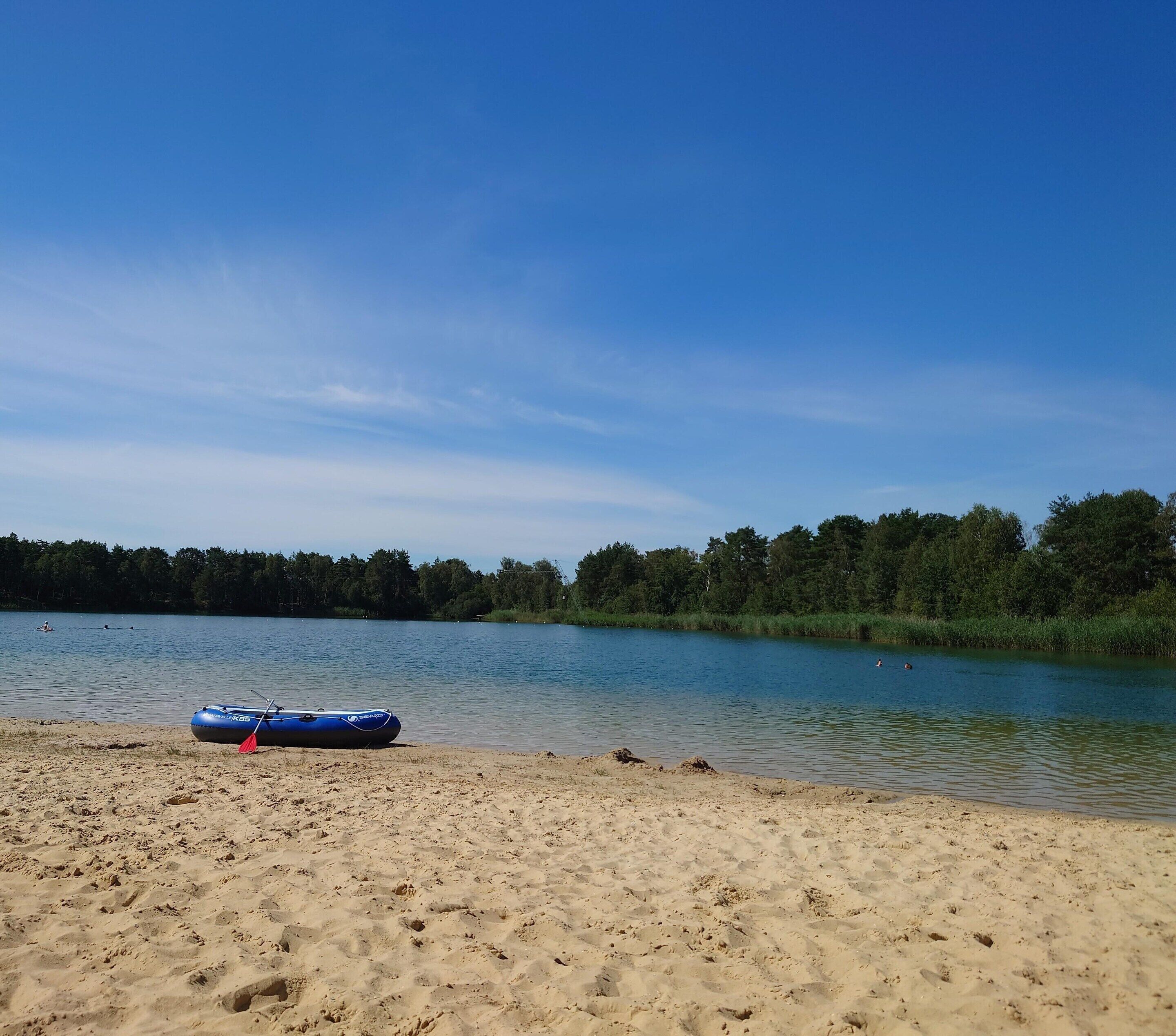 En sandstrand med en blå, uppblåsbar flotte och en stilla sjö.