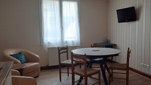 Ein Zimmer mit einem runden Tisch, zwei Holzstühlen, einem Sofa mit blauem Kissen, einer kleinen Topfpflanze und einem an der Wand befestigten Fernseher.