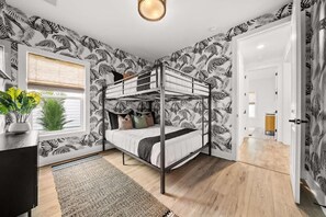 Ein Zimmer mit einem Etagenbett, einem Schreibtisch mit Pflanze, einem Fenster mit Jalousien und einer Tür zu einem weiteren Raum.