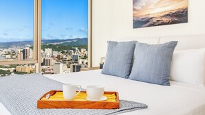 A hotel room with a large window offering a city view, a bed with white bedding, two blue pillows, and a tray with two white cups on a wooden tray. - Luxury Sky Ala Moana Condo with Ocean & City Views (Honolulu)