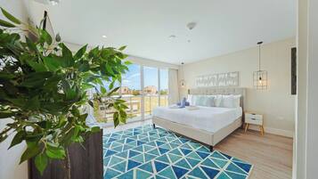 Une chambre moderne avec un grand lit, un tapis bleu aux motifs géométriques, une plante en pot et une vue sur la ville grâce à de grandes fenêtres.