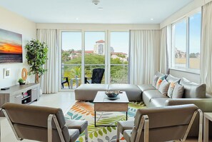 A modern living room with a sofa, chairs, a coffee table, and a large window with a view of buildings and greenery. - Palm Beach Condo 3 Min. Walk 2 Beach & Restaurants (Noord)