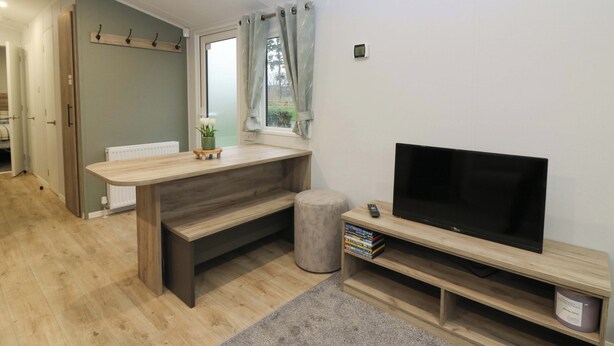 A modern living room with a wooden bench, a TV stand with a flat-screen TV, and a small bookshelf. - 63 St Andrews (Prestonpans)