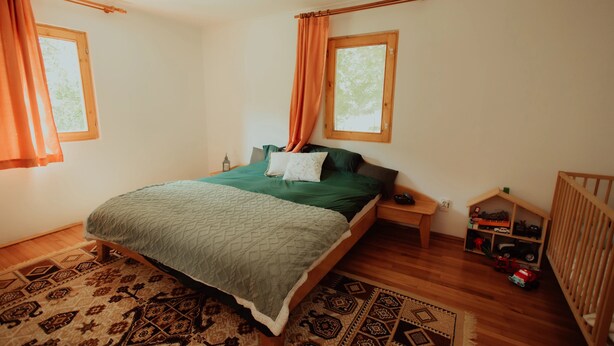 Un dormitorio con cama, suelo de madera, alfombra estampada, una mesita de noche de madera, una cuna y una ventana con cortinas.