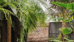A narrow pathway leading to a wooden door with a thatched roof, surrounded by lush greenery and bamboo structures. - LIA Retreat Ba Na Hills (Da Nang)
