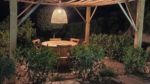 En utomhuspergola av trä med ett runt bord och stolar, omgiven av grönska.