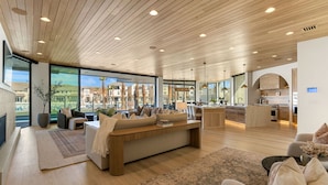 A spacious living area with a wooden ceiling, large windows, and a modern kitchen in the background. - The Oasis Utah: Lagoon-Front, Pool, Slide, Theater (St. George)