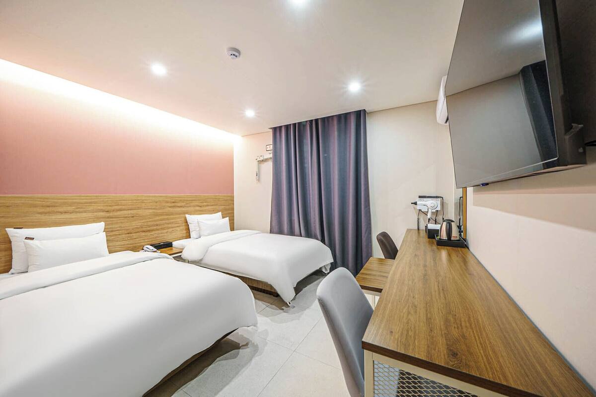 Ein Hotelzimmer mit zwei Betten, einem Holztisch, einem Fernseher und einem Fenster mit Vorhängen.