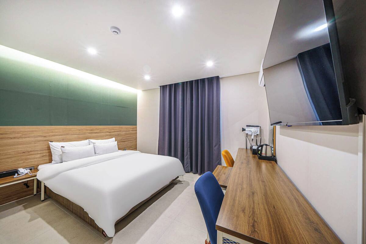 Ein Hotelzimmer mit einem großen Bett, einem Schreibtisch mit Stuhl, einem Flachbildfernseher und einem Fenster mit Vorhängen.