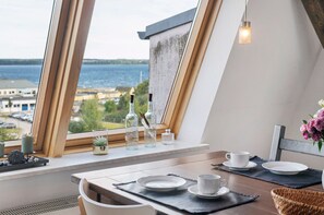 A dining area with a view of the sea, a table set with plates and cups, and a window with bottles and a vase on the sill. - Apartment 'Mit Seeblick' with Lake View and Wi-Fi (Schwerin)