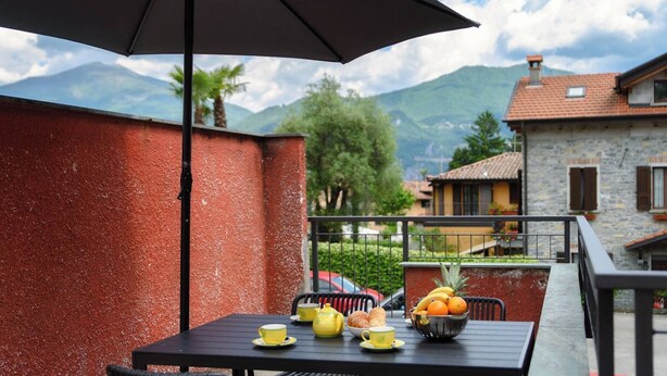 A balcony with a table set for tea, a fruit bowl, and a view of mountains and houses. - Lake Como Retreat - Menaggio (Menaggio)