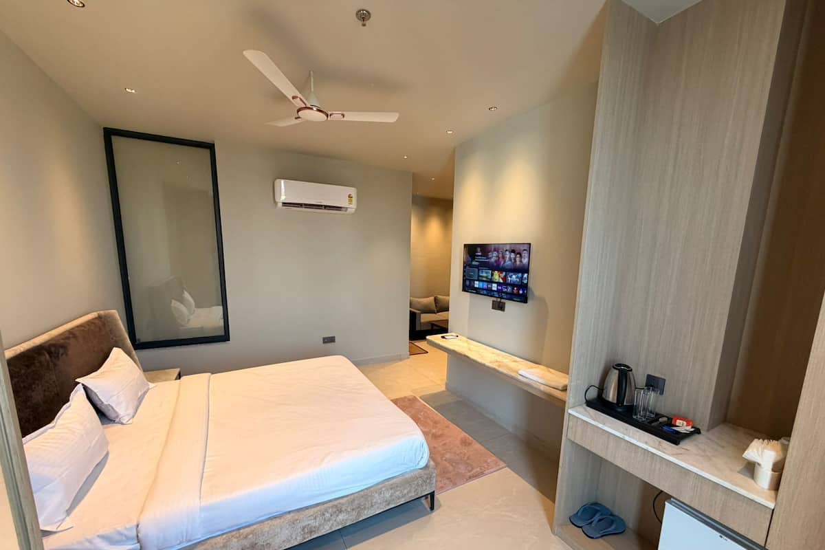 A hotel room with a bed, a TV mounted on the wall, a ceiling fan, and a small desk with a refrigerator.