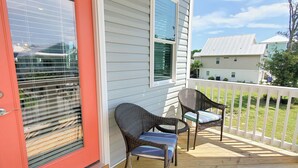 Un porche abrité avec deux fauteuils en osier et une table, une porte rouge et une vue sur les maisons voisines.