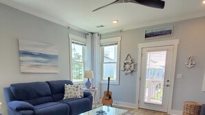 Un salon avec un canapé bleu, une table basse en verre, une guitare et une vue sur l’extérieur, visible à travers une porte vitrée.