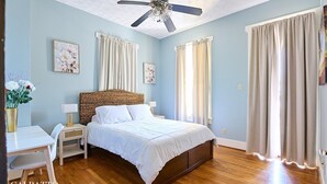 Un dormitorio con cama, escritorio, silla, ventilador de techo y cortinas.