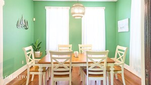 Comedor con mesa de madera, sillas blancas, paredes verdes y una lámpara colgante.