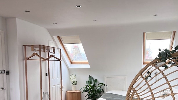 A modern bedroom with a skylight, a bed with a green blanket, a rattan chair, and a wooden dresser. - Zozy Gladstone (London)